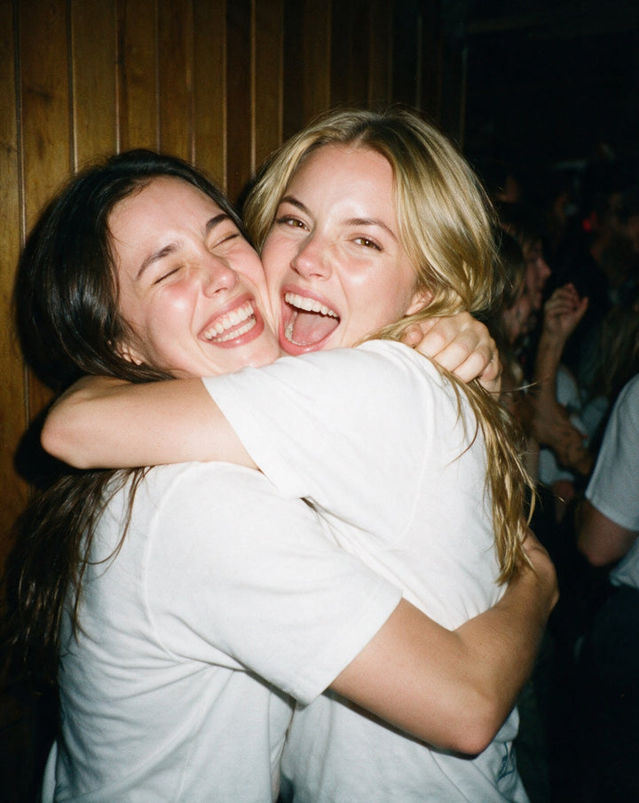 Two friends laughing in a hug at a party, both in white TipsyTees tees